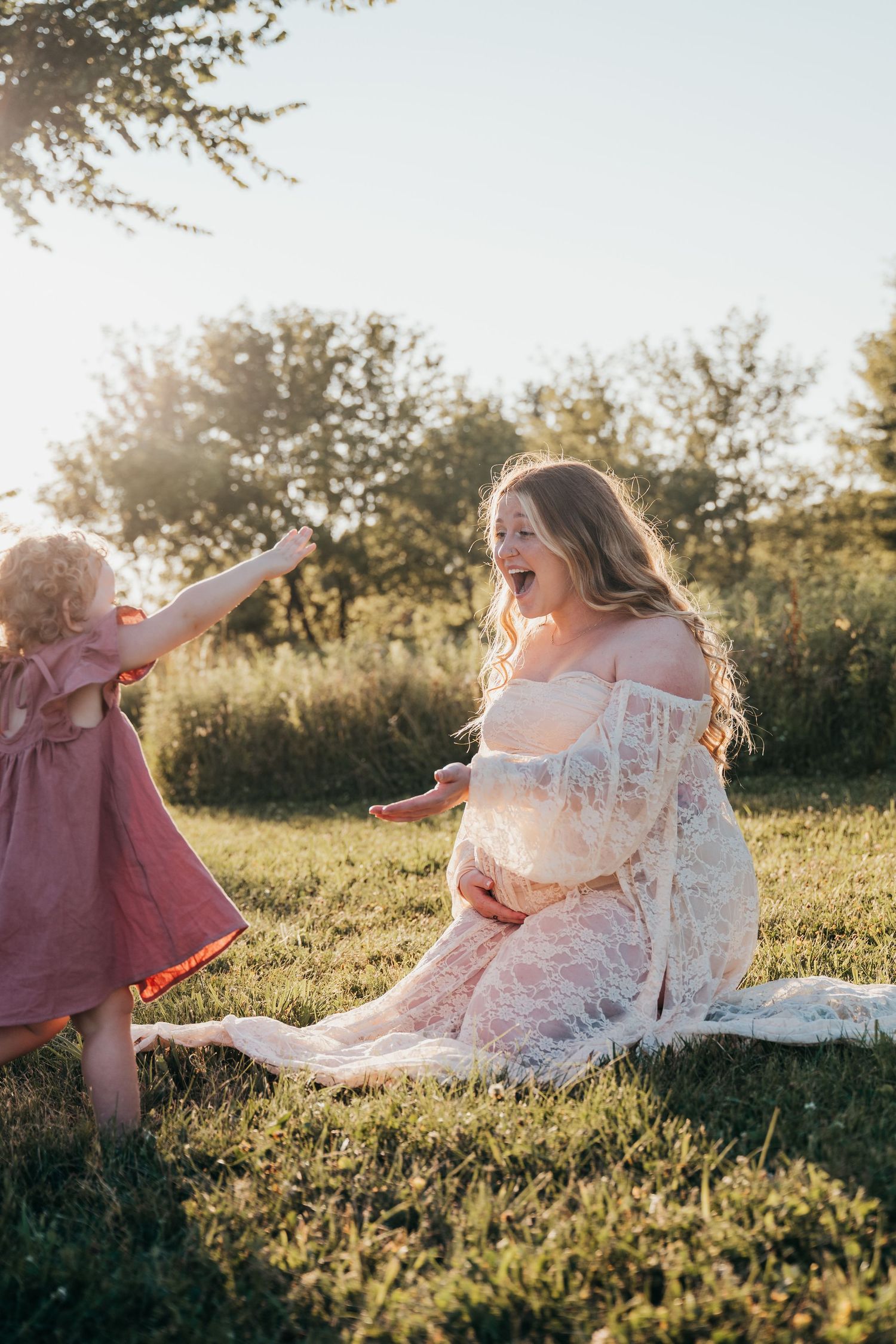 A playful outdoor maternity photo showing a mother in a lace dress sharing a moment with a toddler.