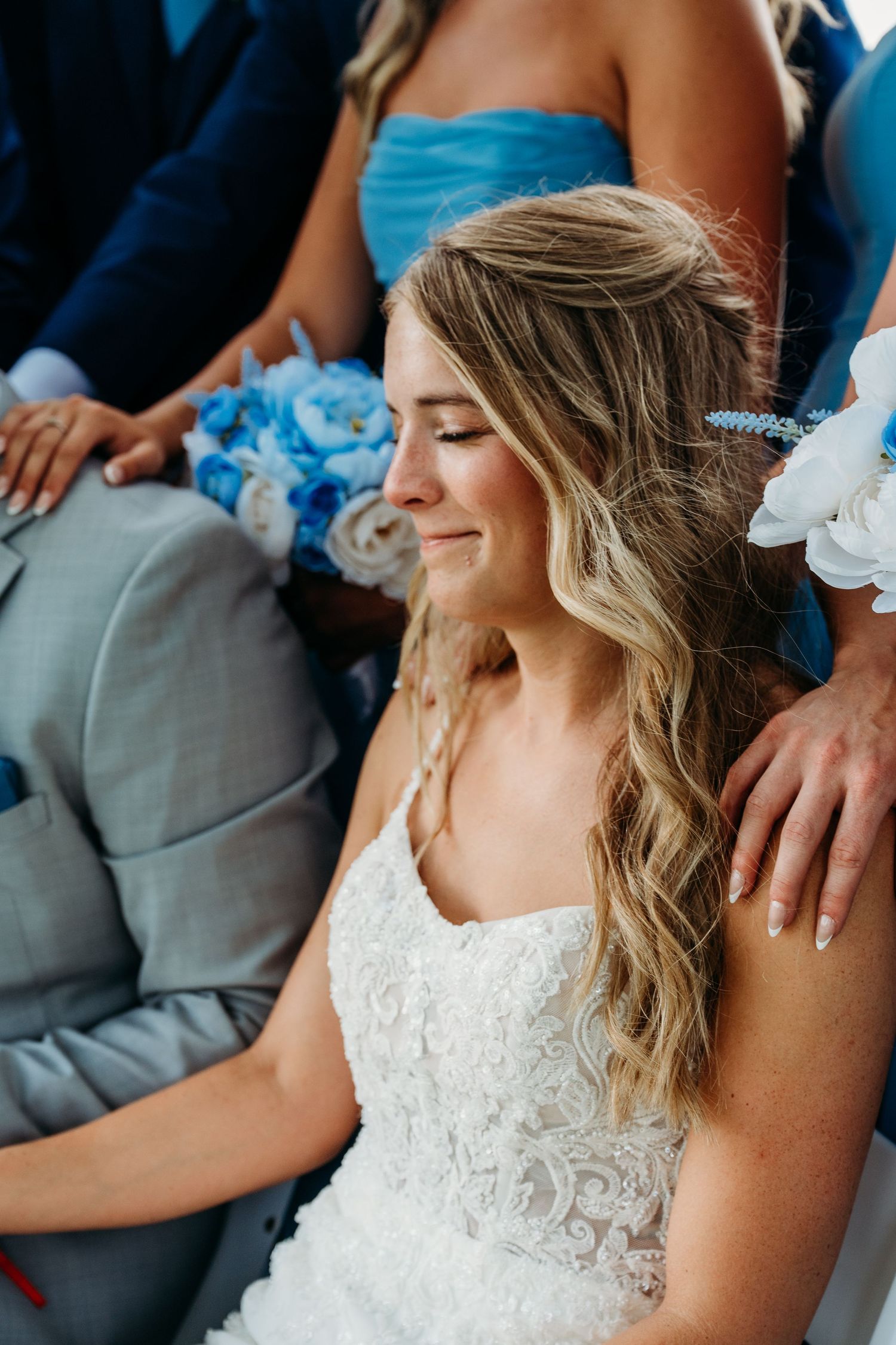 A bride in a white lace wedding dress sits with guests in formal attire at a celebration.