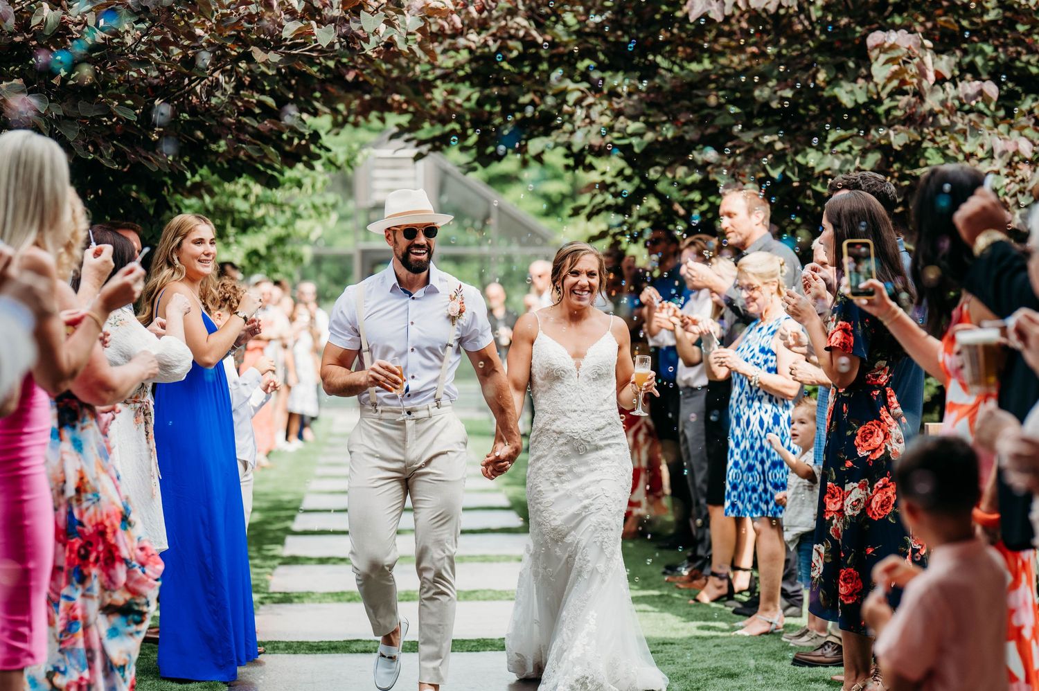 Wedding guests line pathway outdoors cheering and celebrating newlyweds during recessional.