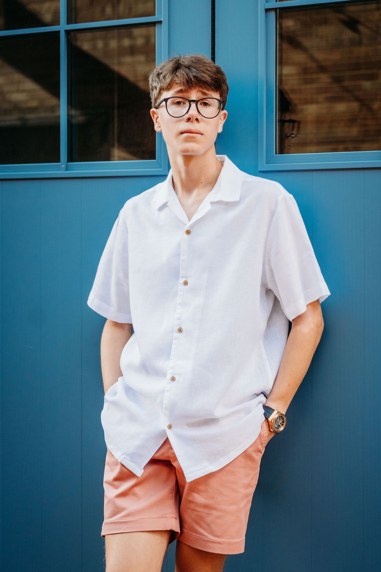 Someone in a white shirt and coral shorts poses against a blue wall outdoors.