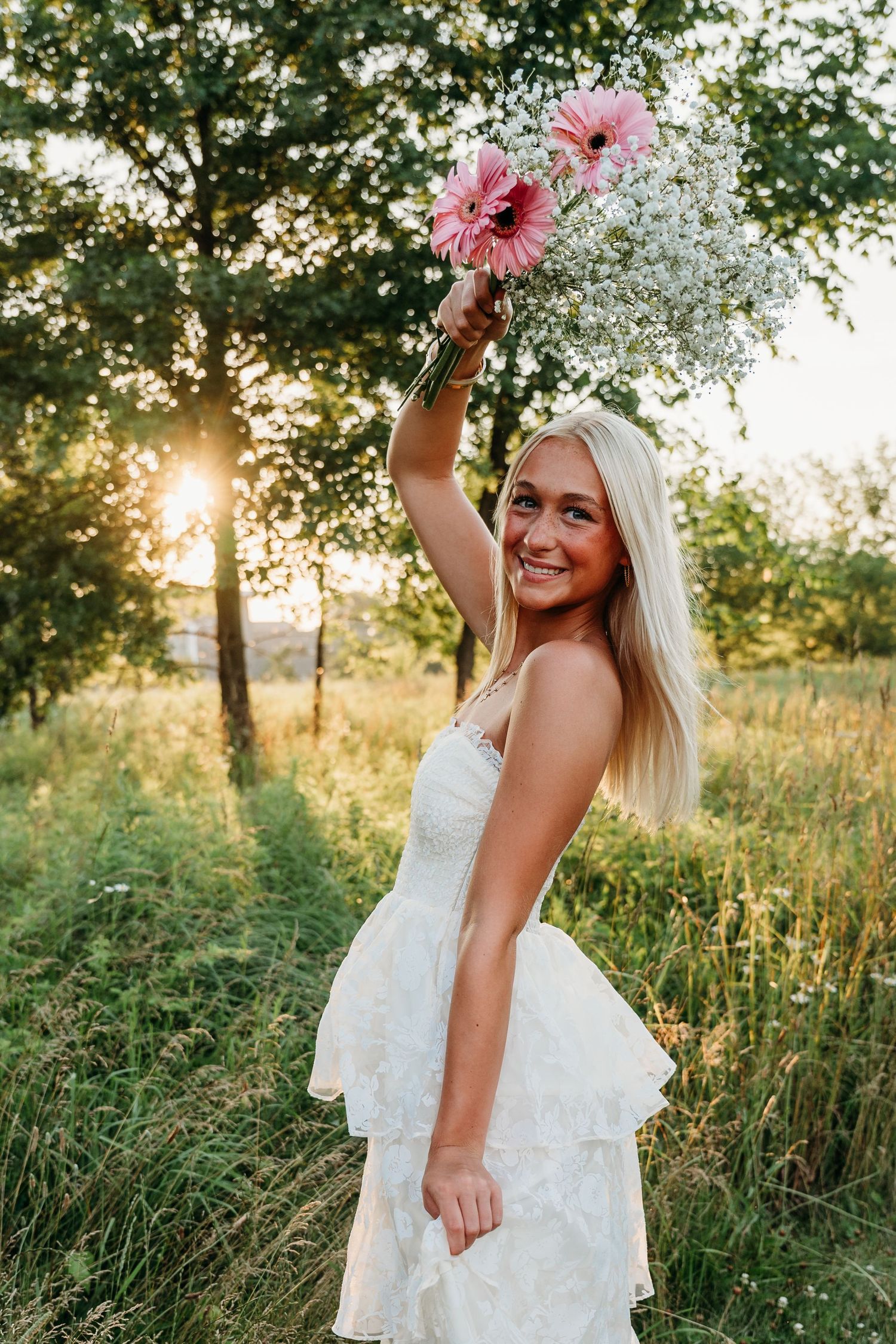 A romantic outdoor photoshoot in a field at sunset features a white lace dress and pink gerbera daisies.