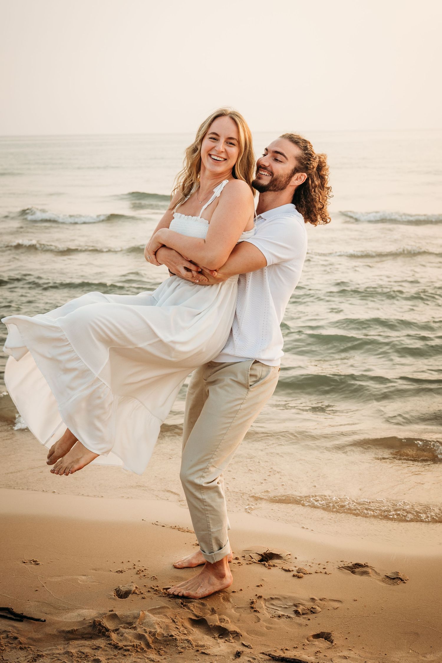 A joyful couple embraces on a sandy beach at sunset, man lifting woman in white dress with ocean waves behind them.