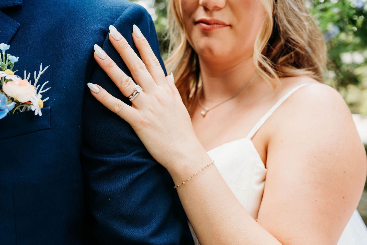 Bride resting hand on groom's navy suit, showcasing wedding ring set, bracelet, and manicured nails at outdoor ceremony.