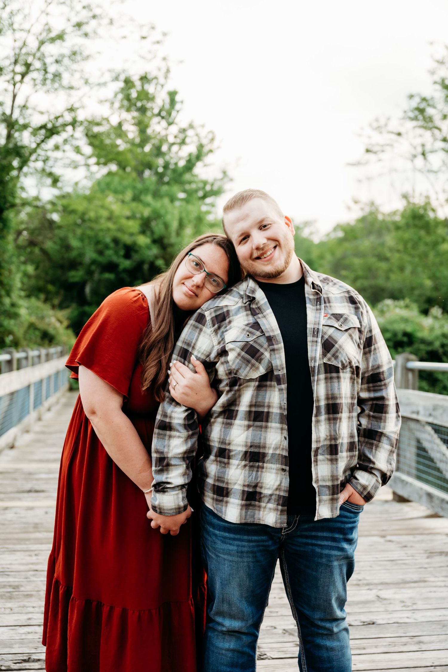 A couple poses on a wooden bridge, woman in red dress leaning on man in plaid shirt and jeans, surrounded by green trees.