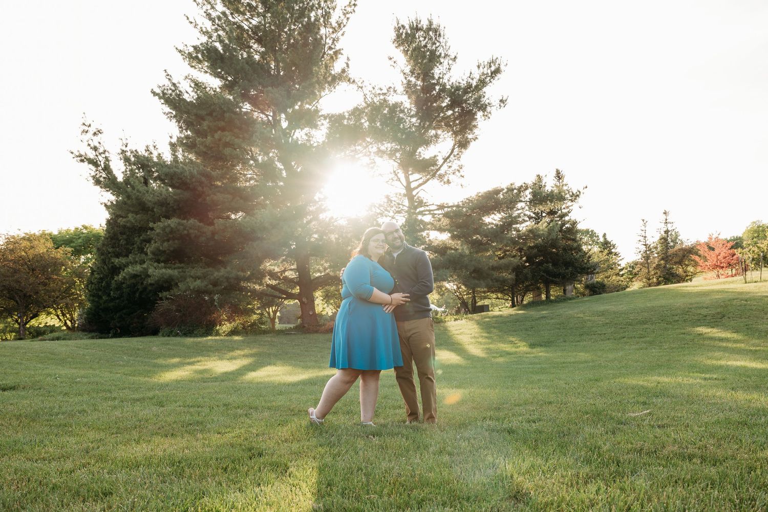 Couple embracing in a sunlit green park, golden hour light streaming through tall trees, woman in blue dress.