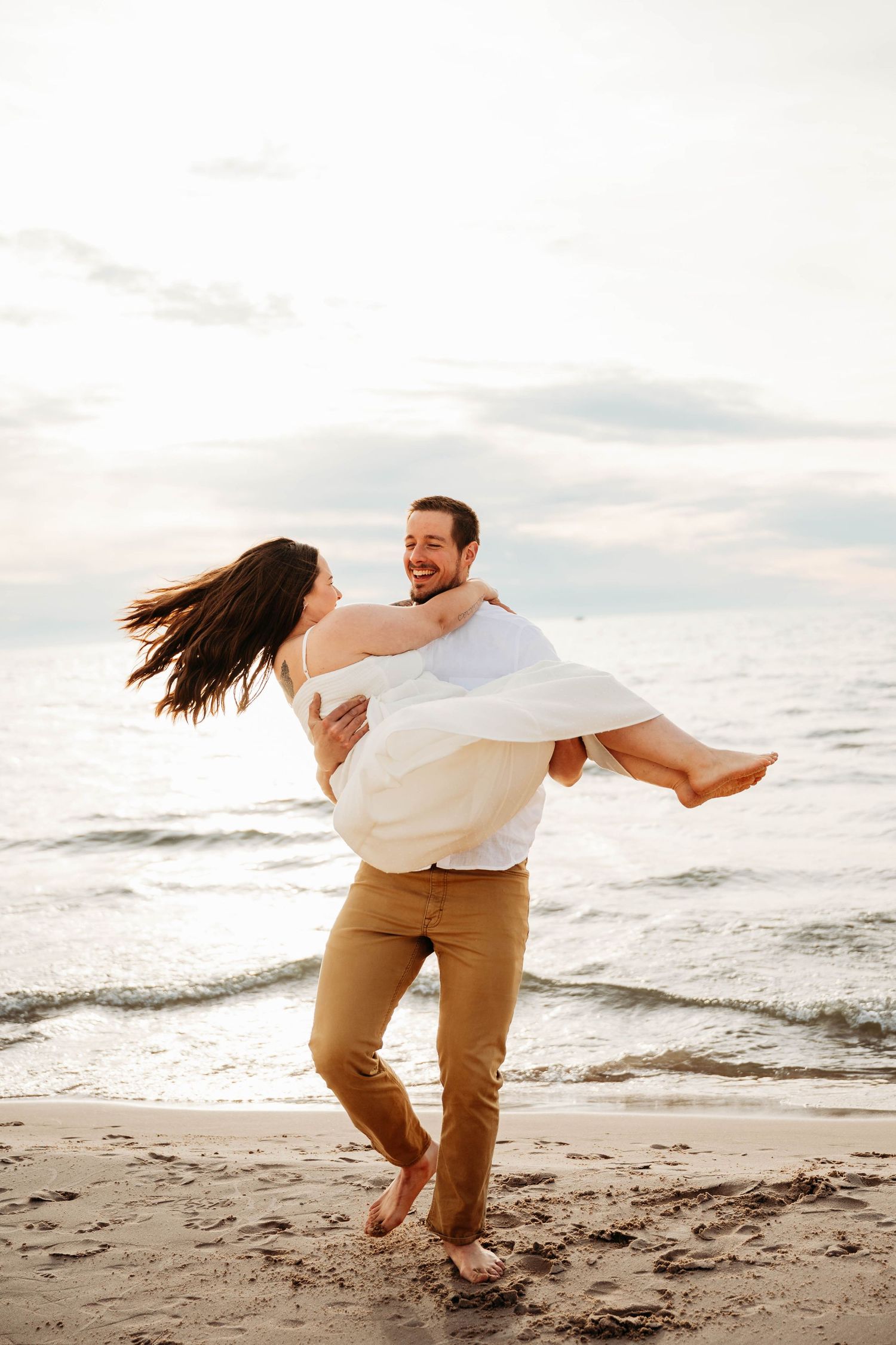 Man carrying woman in white dress on beach at golden hour, both laughing joyfully.
