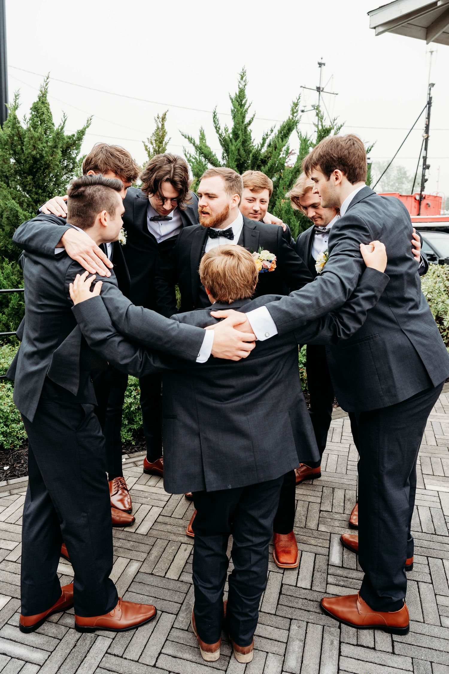Groom and groomsmen in black suits huddle together in celebratory group embrace on wedding day.