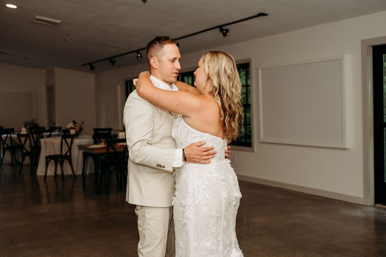 Couple shares tender moment during their first dance in spacious reception venue with white walls and track lighting.
