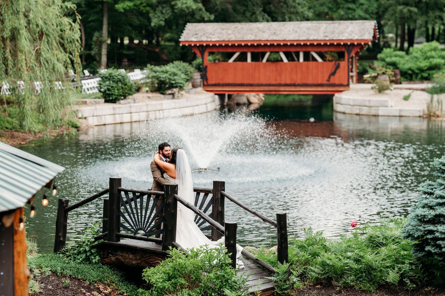 Bride and groom embrace on wooden bridge by fountain with red covered bridge backdrop at scenic outdoor wedding venue.