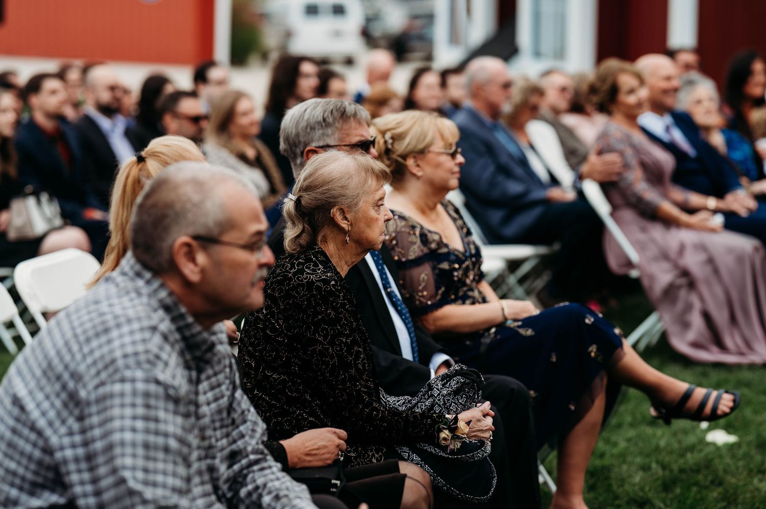 Wedding guests sit in rows during an outdoor ceremony on a lawn with trees in the background.