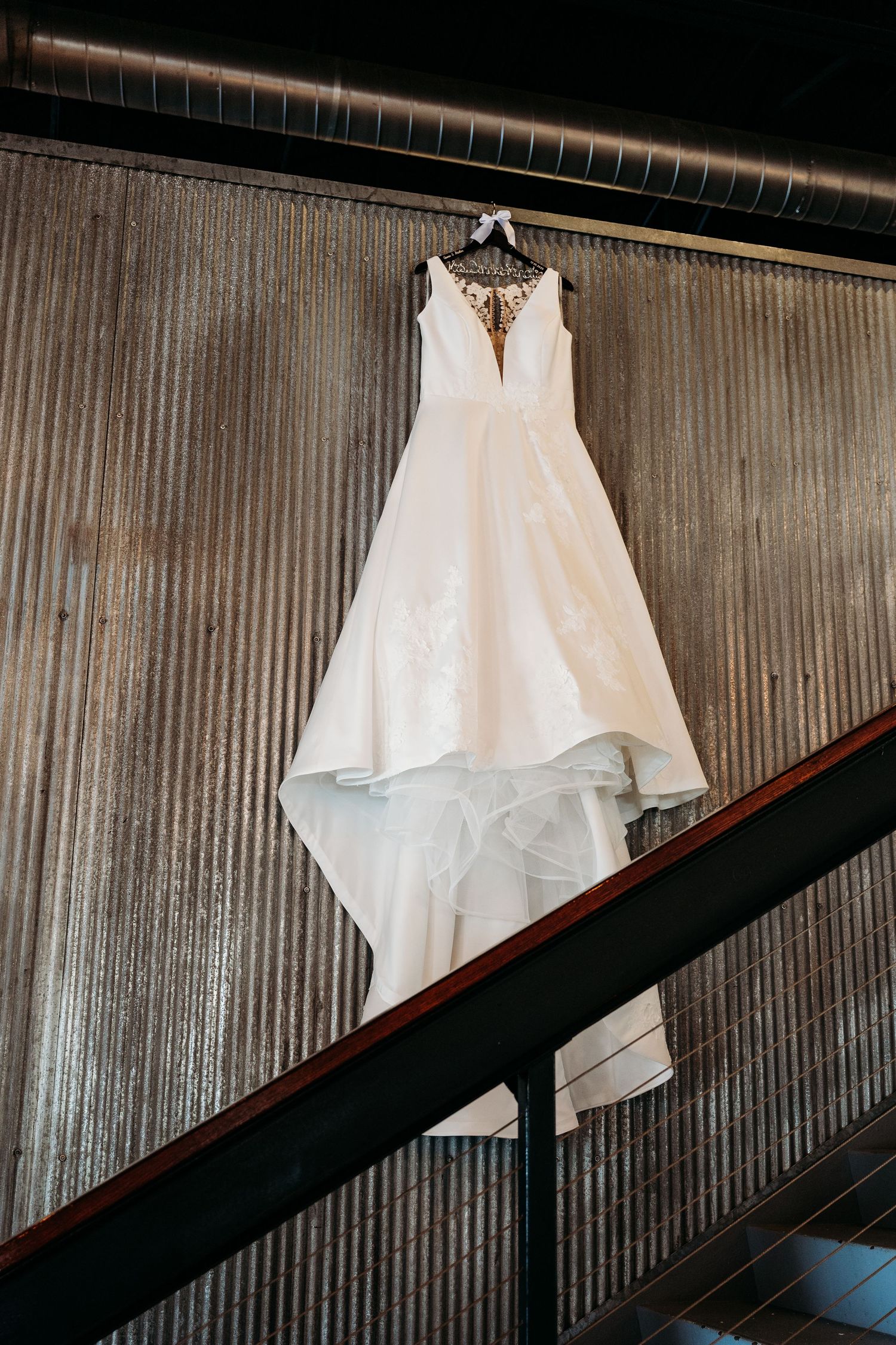 Elegant white wedding dress hangs on display against a textured metallic wall with dark railings.