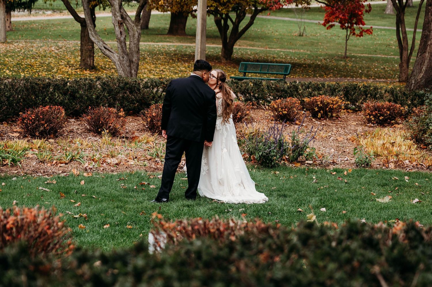 Romantic autumn wedding couple embraces among fall foliage and trees in an outdoor garden setting.