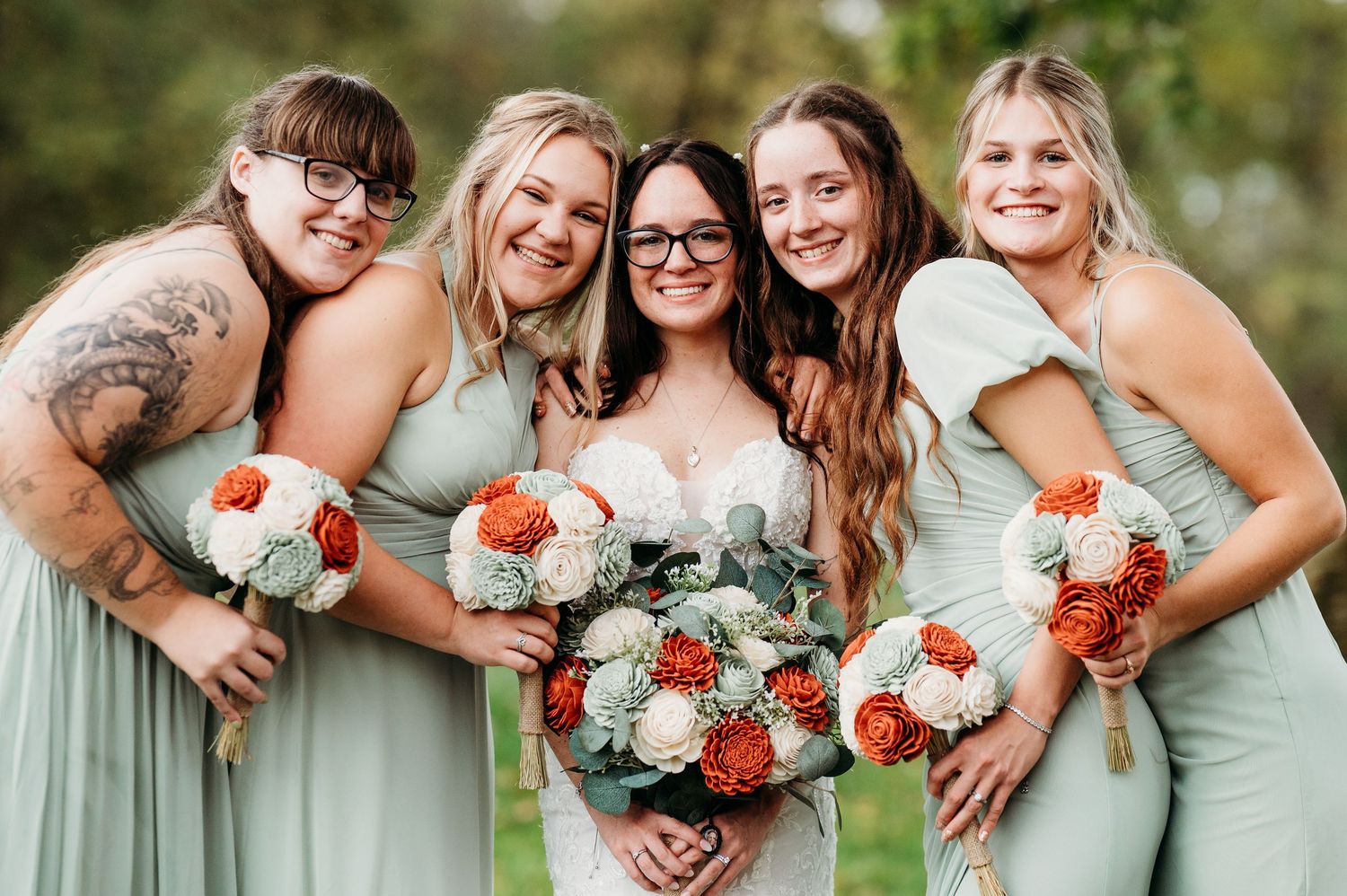 Bridesmaids in mint green dresses pose together with coral and white floral bouquets during an outdoor wedding.