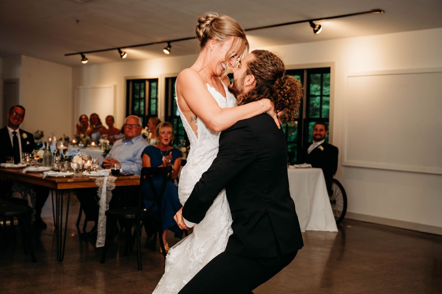 Groom lifts bride during first dance at wedding reception with guests seated at tables in background.