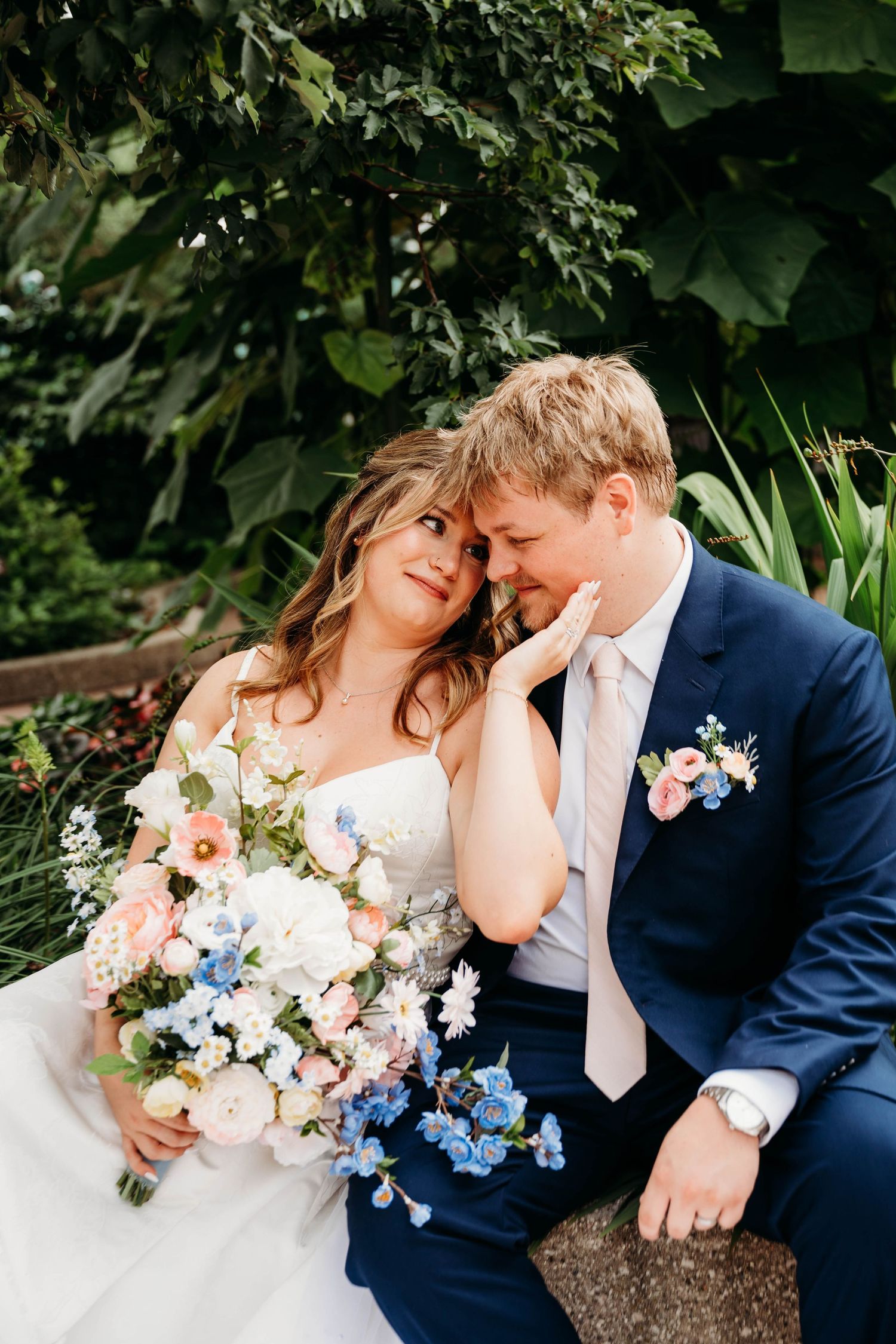 Newlyweds sit together in garden, bride holding pink and blue bouquet as groom tenderly cups her face.