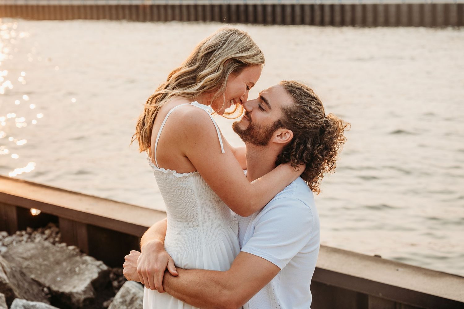 A couple shares a romantic kiss near a sunlit waterfront, the man lifting the woman in a tender embrace.