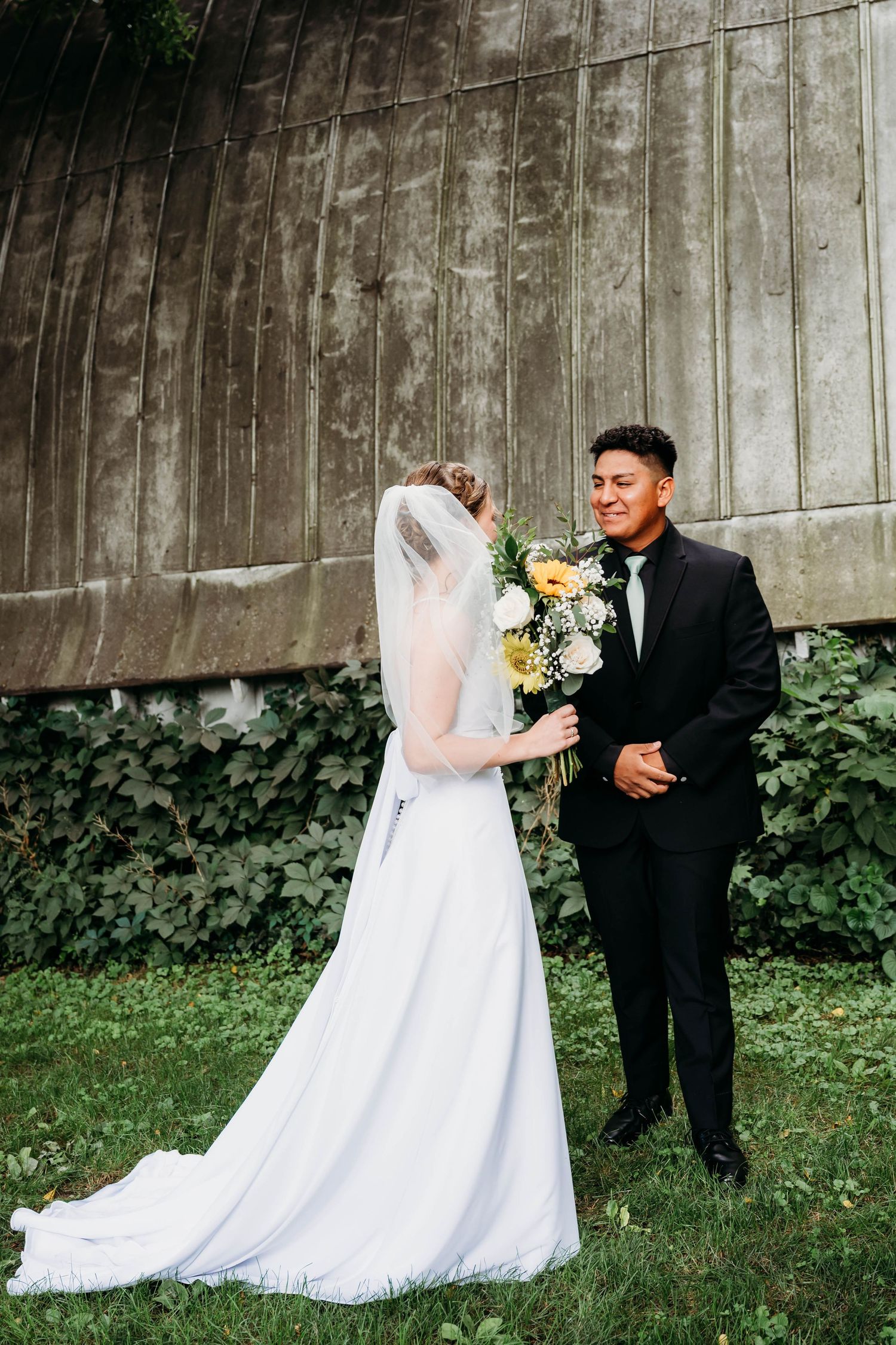 Bride in white gown with veil holds yellow bouquet facing smiling groom in black tuxedo outdoors.