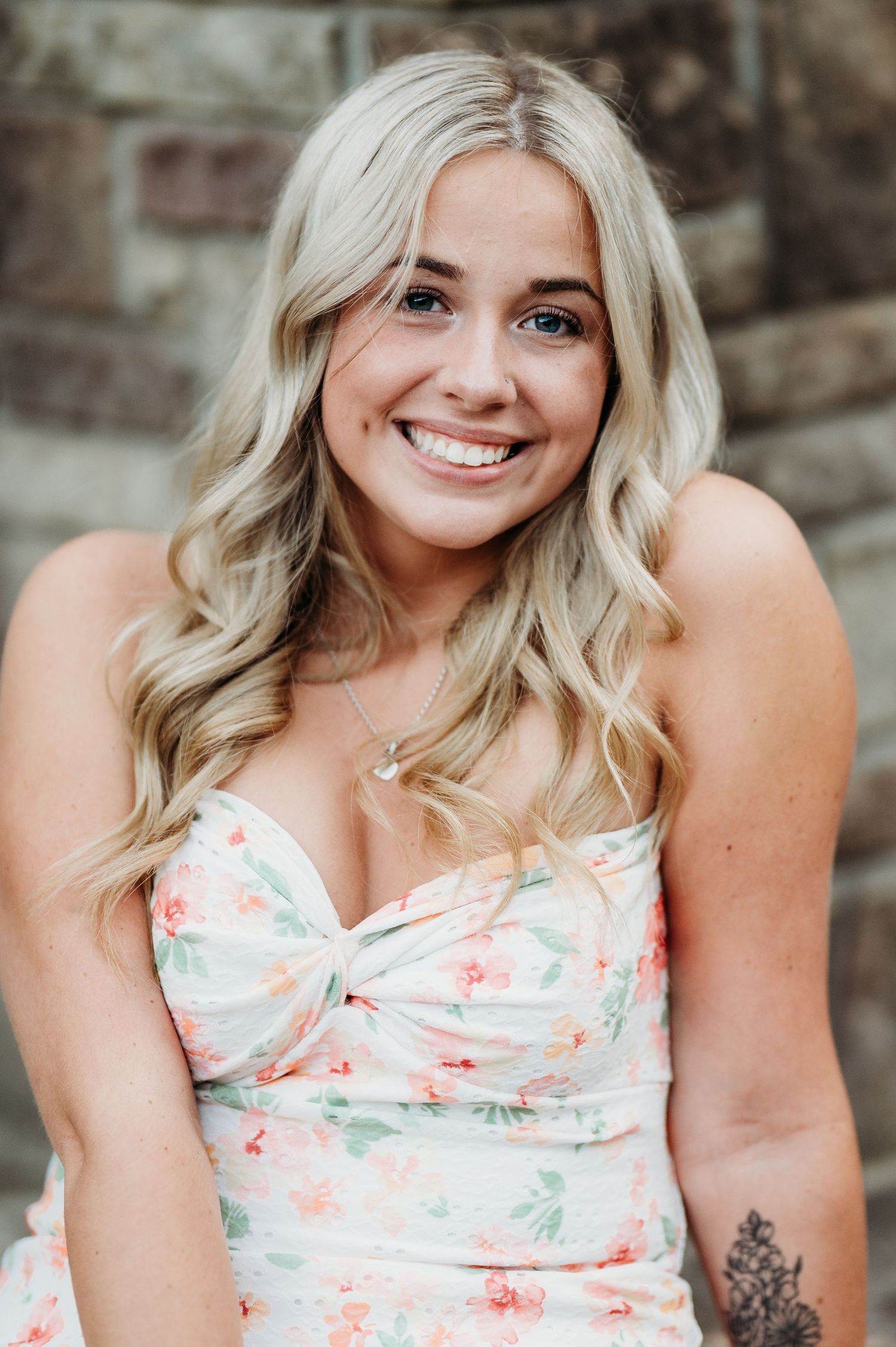 A woman in a floral sundress with blonde wavy hair smiles warmly against a stone wall background.