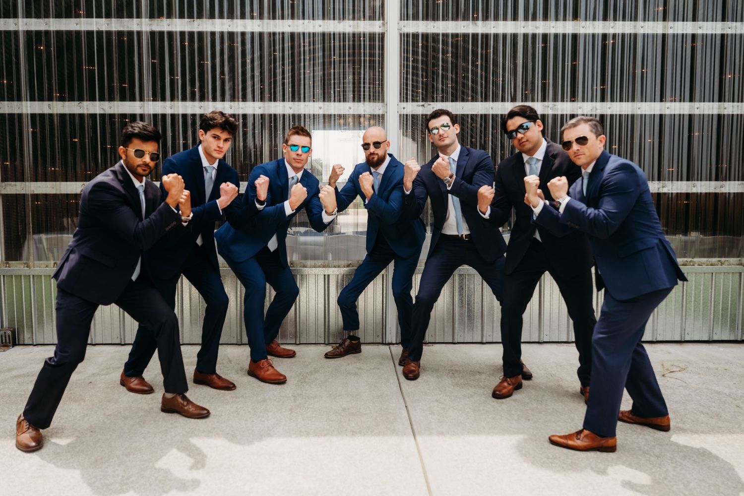Seven groomsmen in navy blue suits and brown shoes pose playfully in fighting stances outdoors at a wedding.