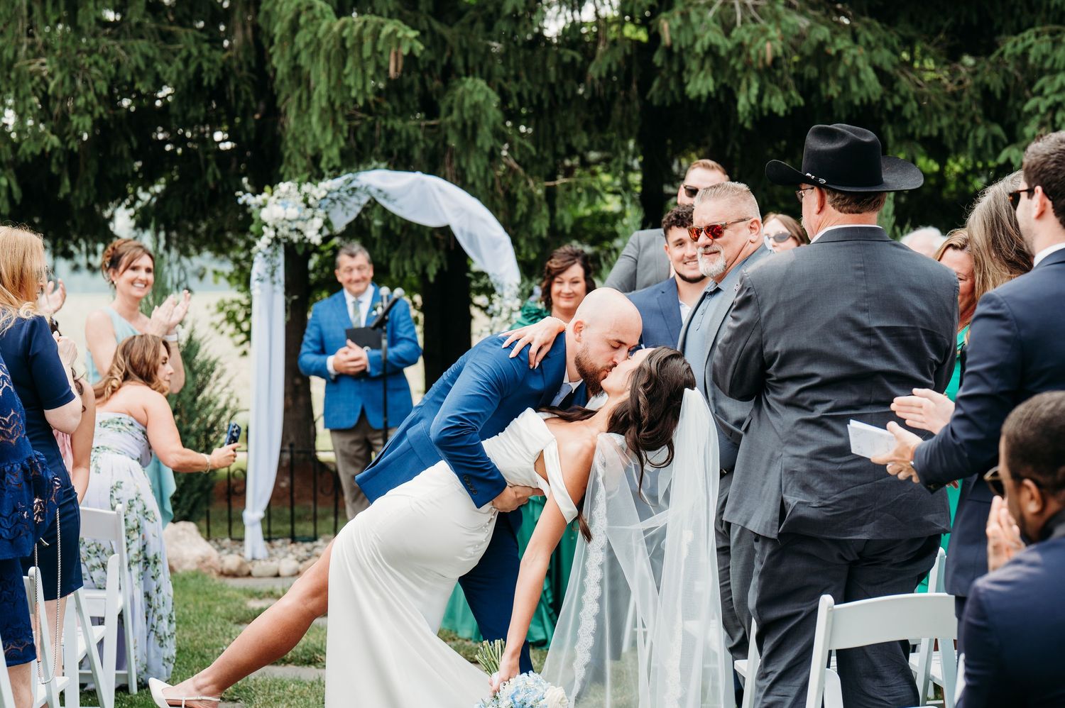 Groom dips bride for a kiss at outdoor wedding ceremony with floral arch and guests celebrating.