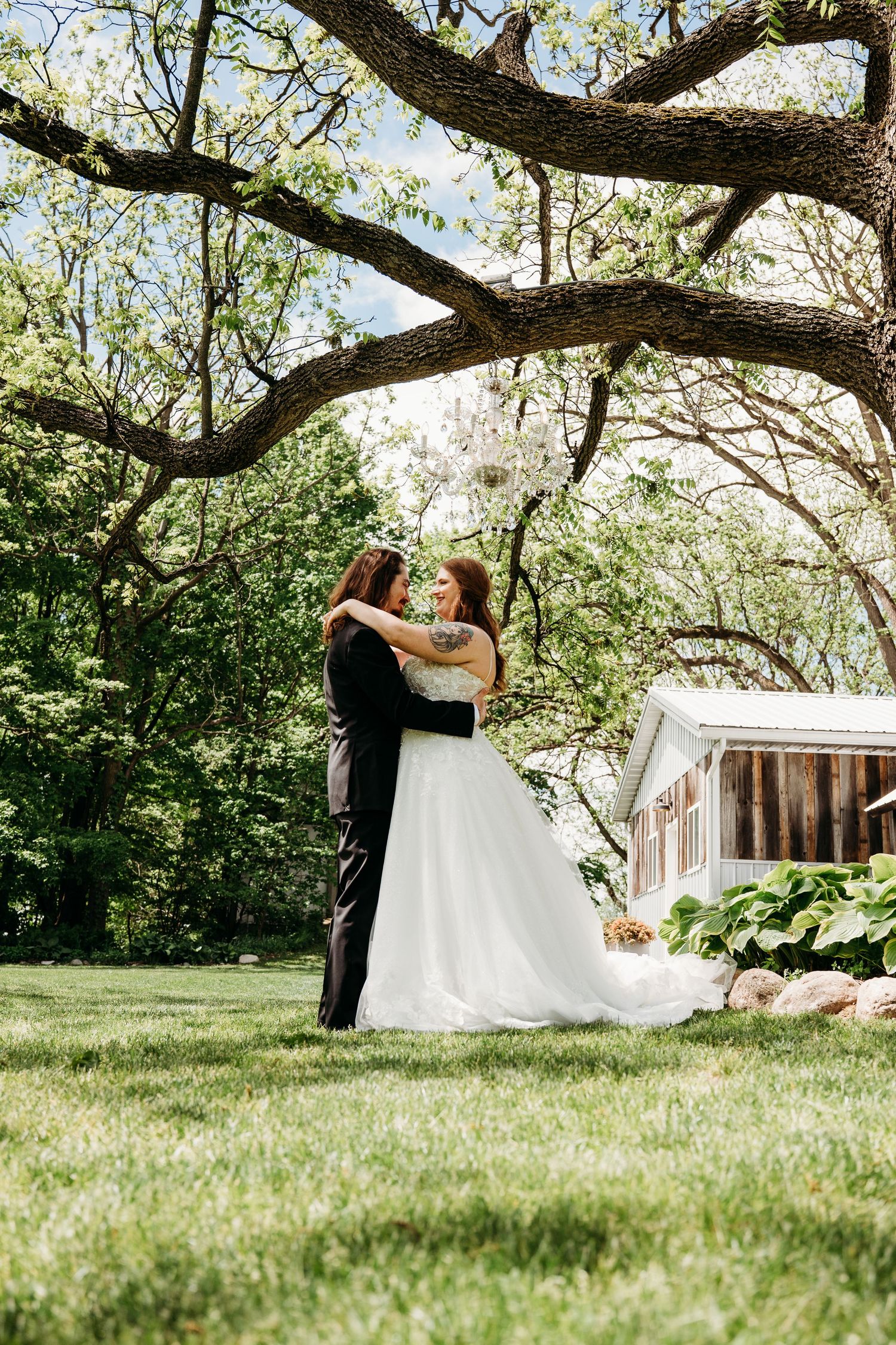 A couple embraces under a large tree branch on lush grass near a rustic wooden barn on their wedding day.