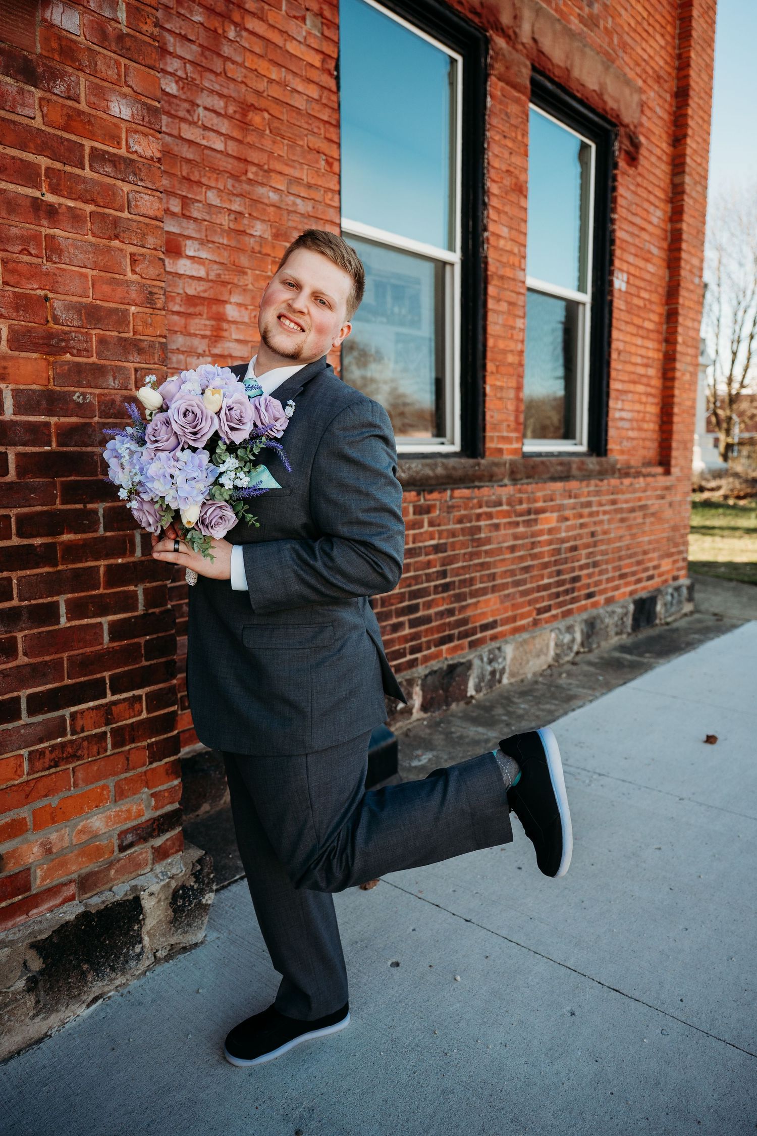 A person in a dark suit poses playfully against a brick wall while holding a purple and lavender floral bouquet.