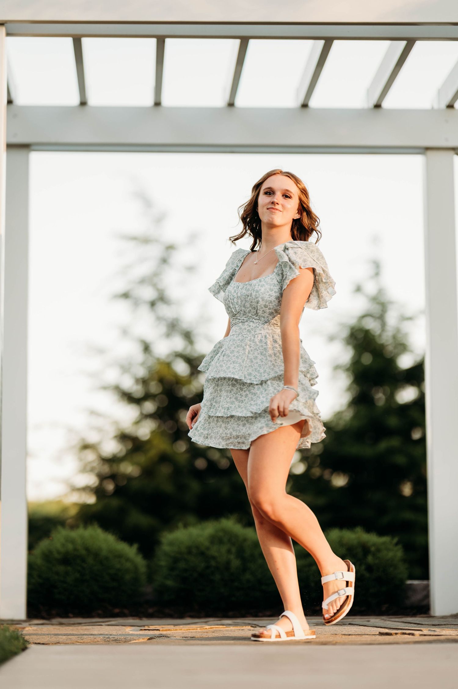 Young woman in floral tiered mini dress and sandals posing under white pergola at golden hour.