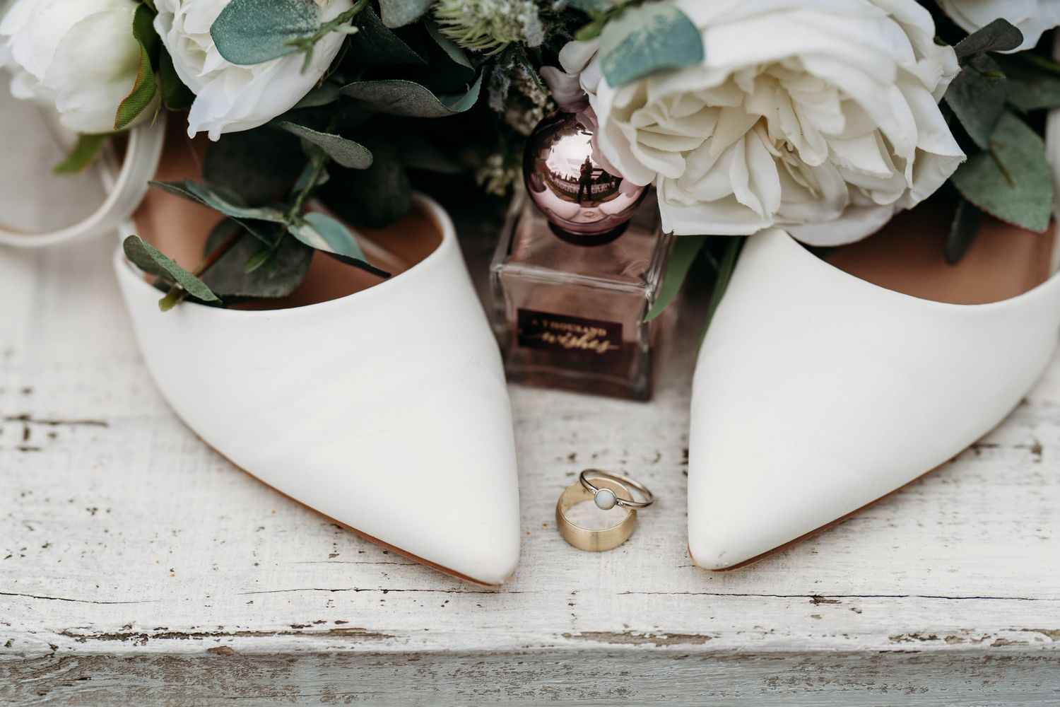 White bridal shoes with elegant floral arrangements and wedding rings styled on a rustic white wooden surface.
