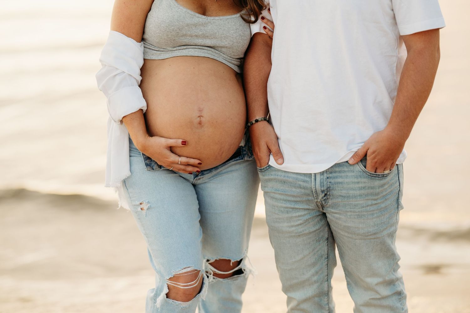 A close up view of a pregnant woman's belly with a couple wearing light denim and white clothing on a beach.