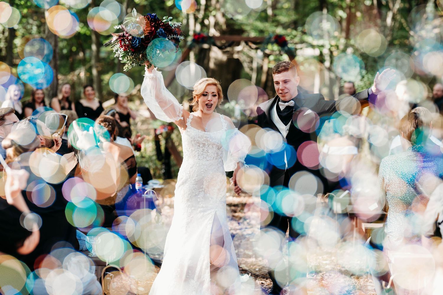 A festive wedding celebration with guests blowing bubbles around people in formal attire during an outdoor ceremony.