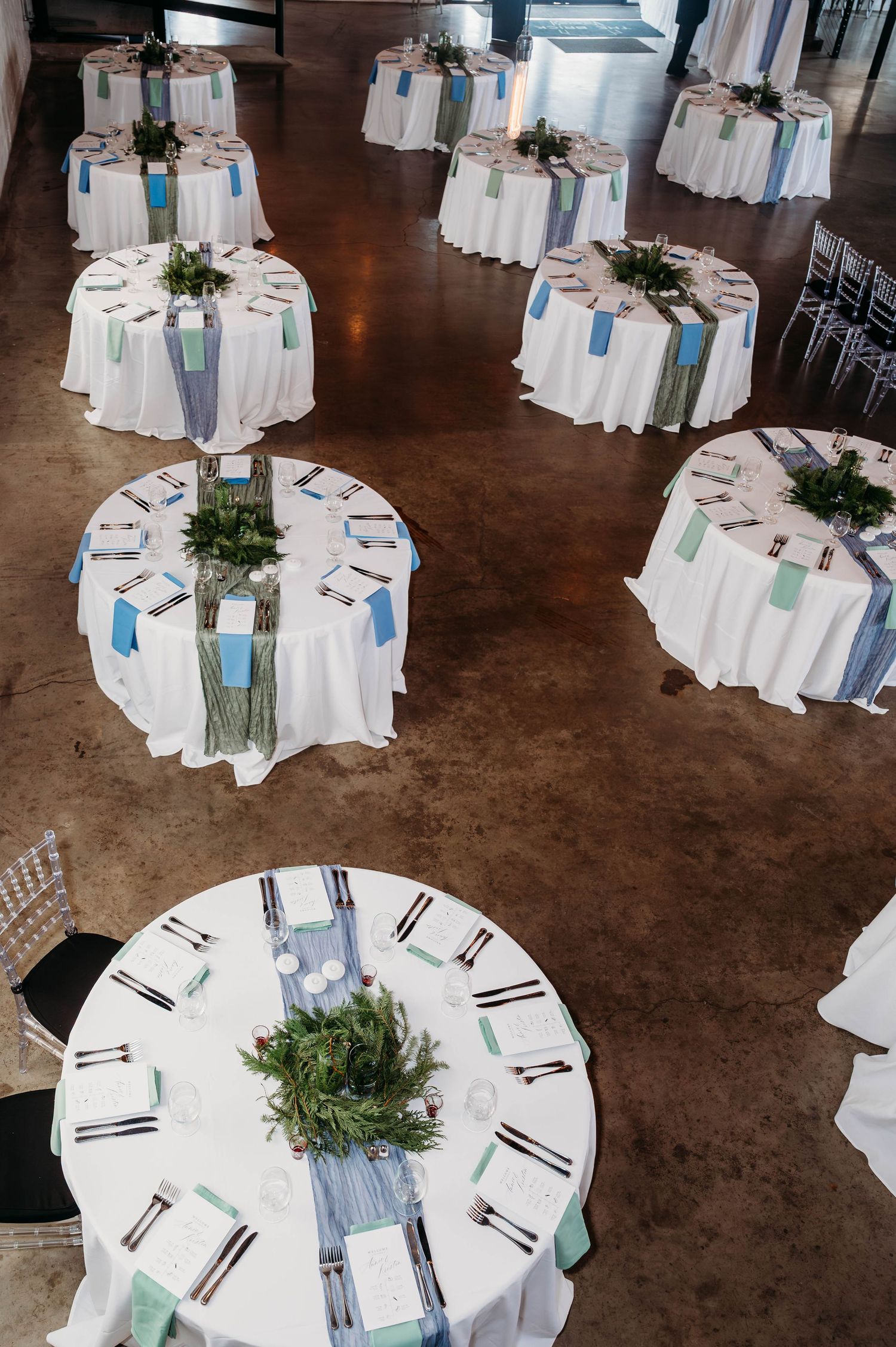 Wedding reception venue with elegant white round tables set with blue and green accents and greenery centerpieces.