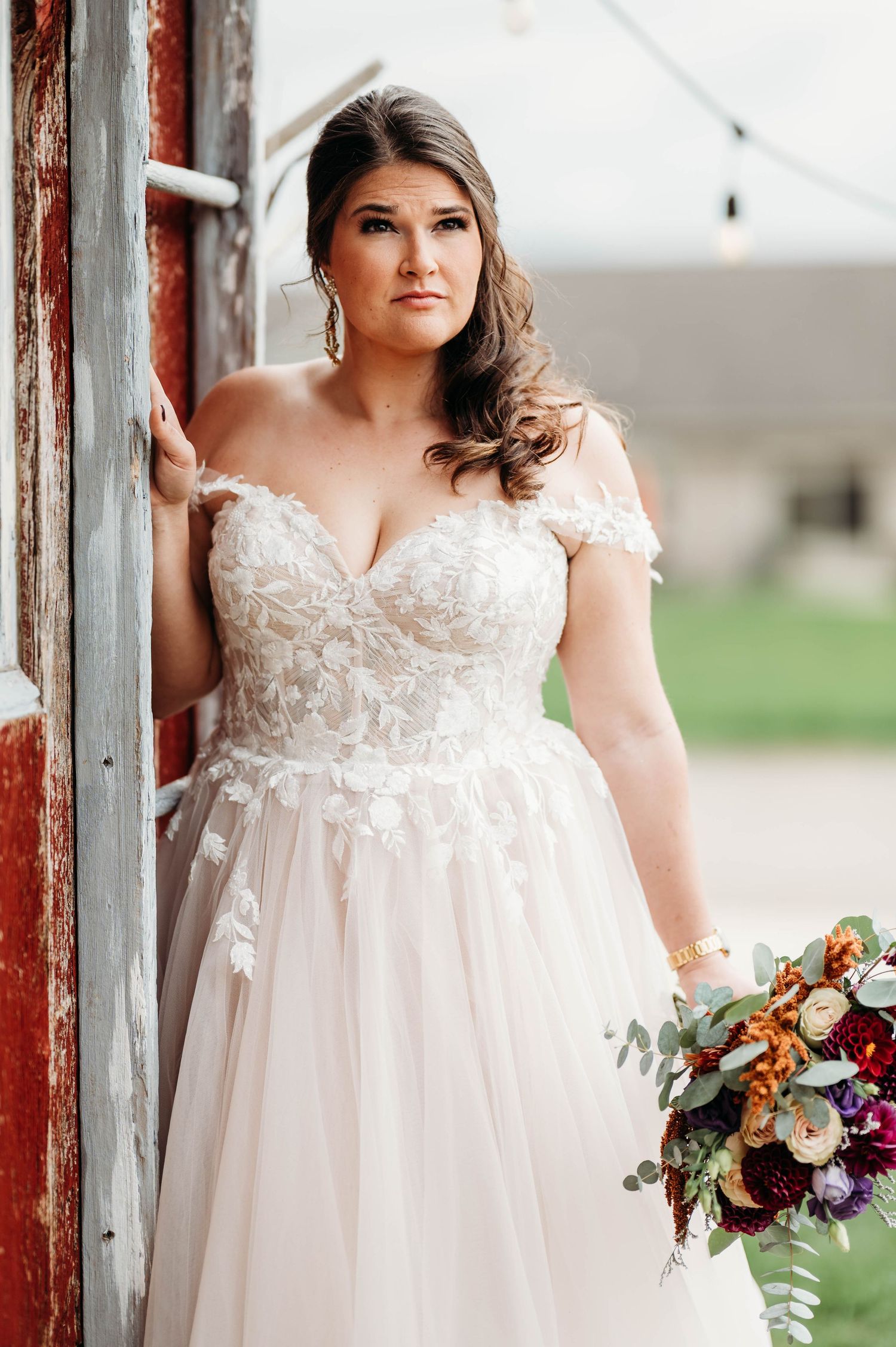 A wedding portrait featuring an elegant lace gown and autumn-toned bouquet against a rustic red barn wall.