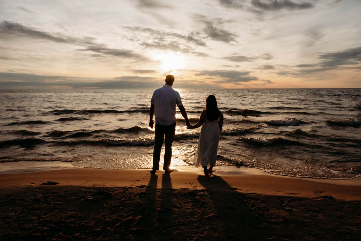 Romantic couple holding hands silhouetted against a stunning ocean sunset on a sandy beach.