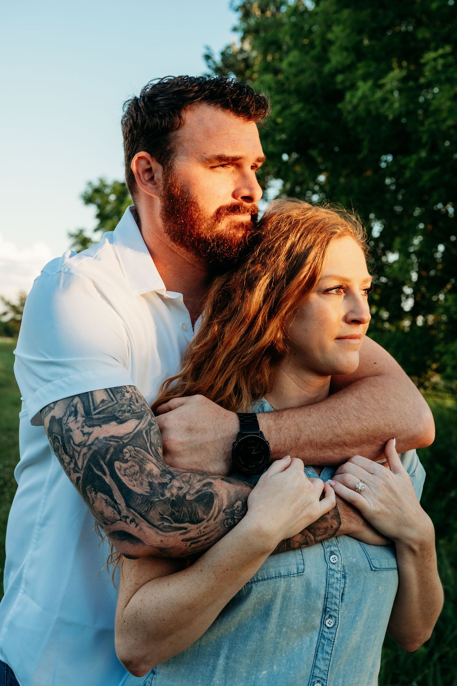 A bearded tattooed man embraces a red-haired woman from behind in a golden hour outdoor portrait.