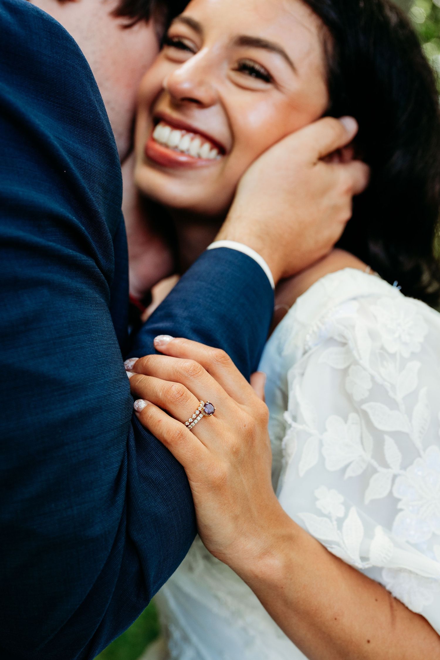 Smiling bride in white lace dress shows off engagement ring while groom in navy suit embraces her face.