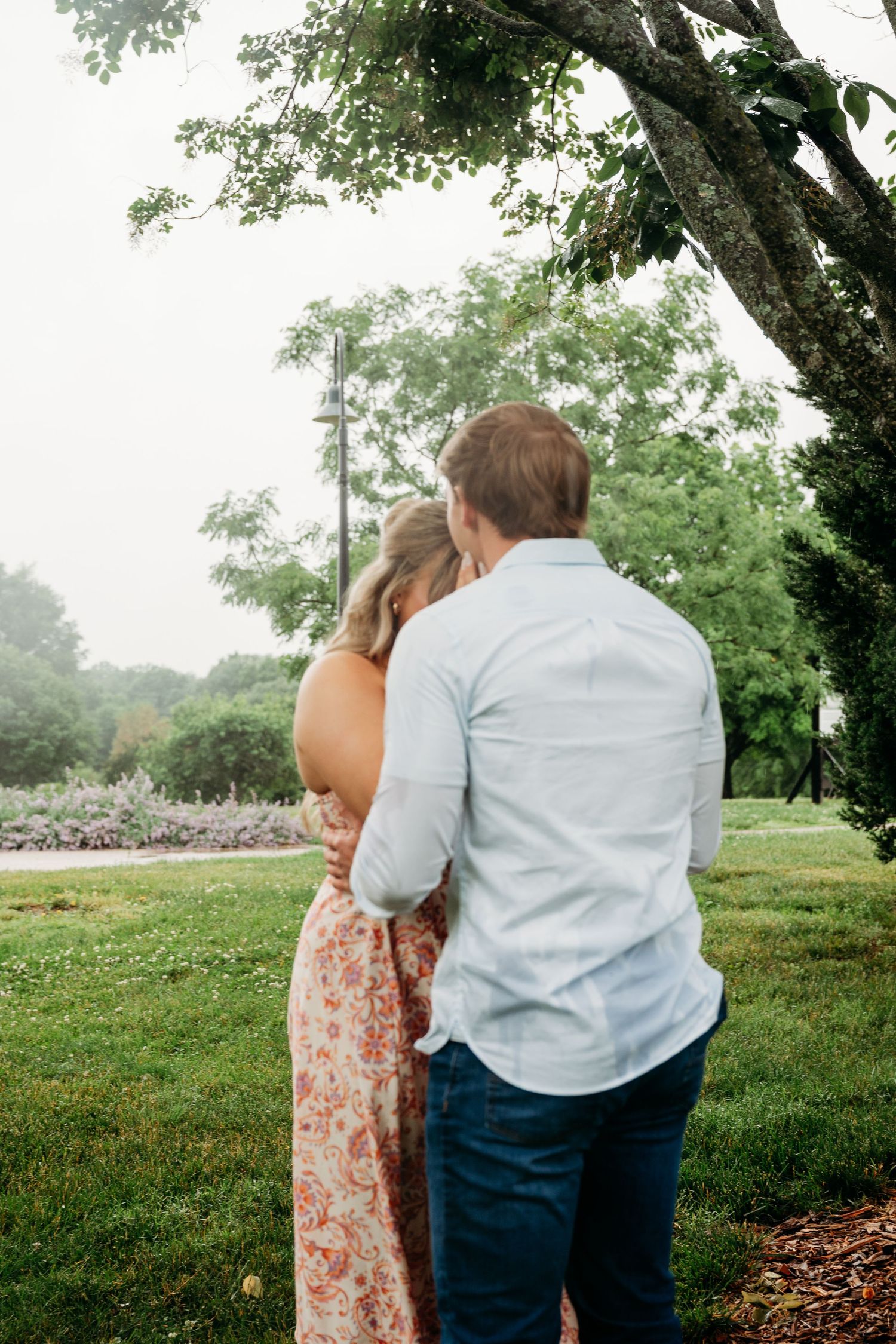 A couple embraces tenderly under a large tree in a lush green park, the woman in a floral dress leaning on the man.