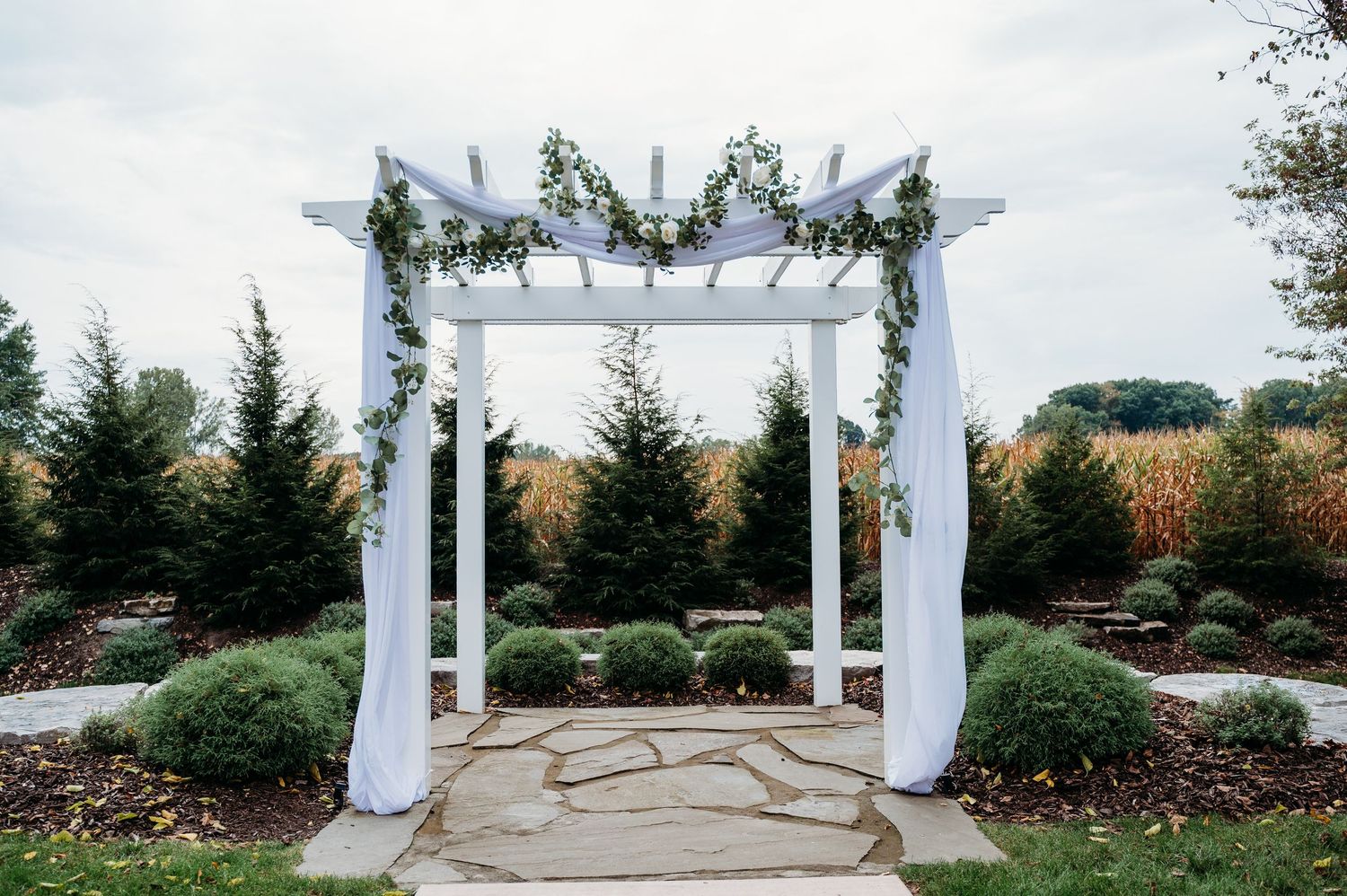 White pergola structure stands elegantly in a garden setting with evergreen trees and landscaping in the background.