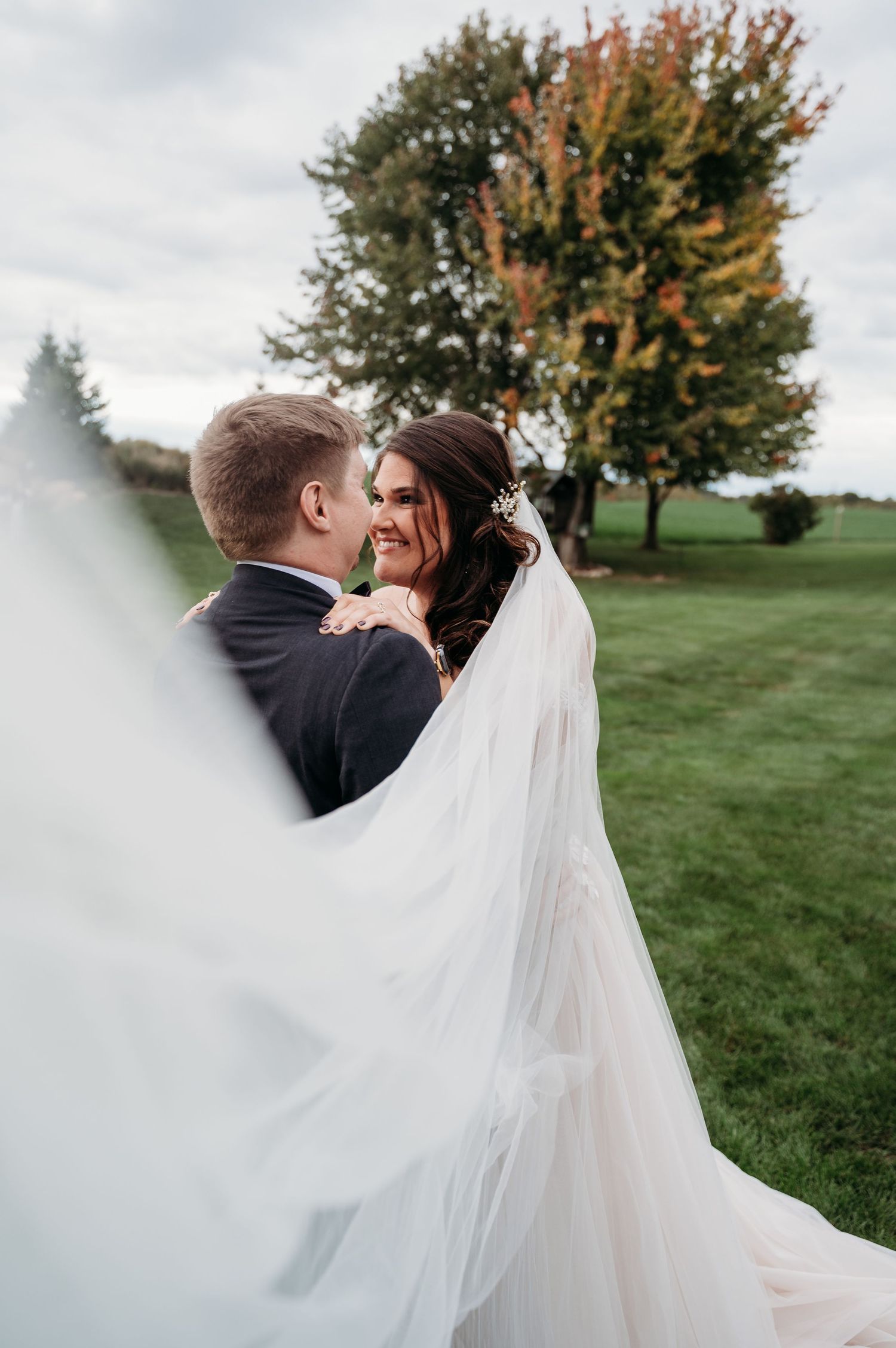 A romantic wedding couple embraces outdoors with a flowing white veil and autumn trees in the background.
