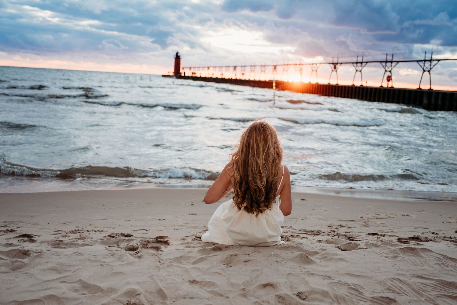 Woman sitting on beach watching waves crash against pier during vibrant sunset over Lake Michigan.