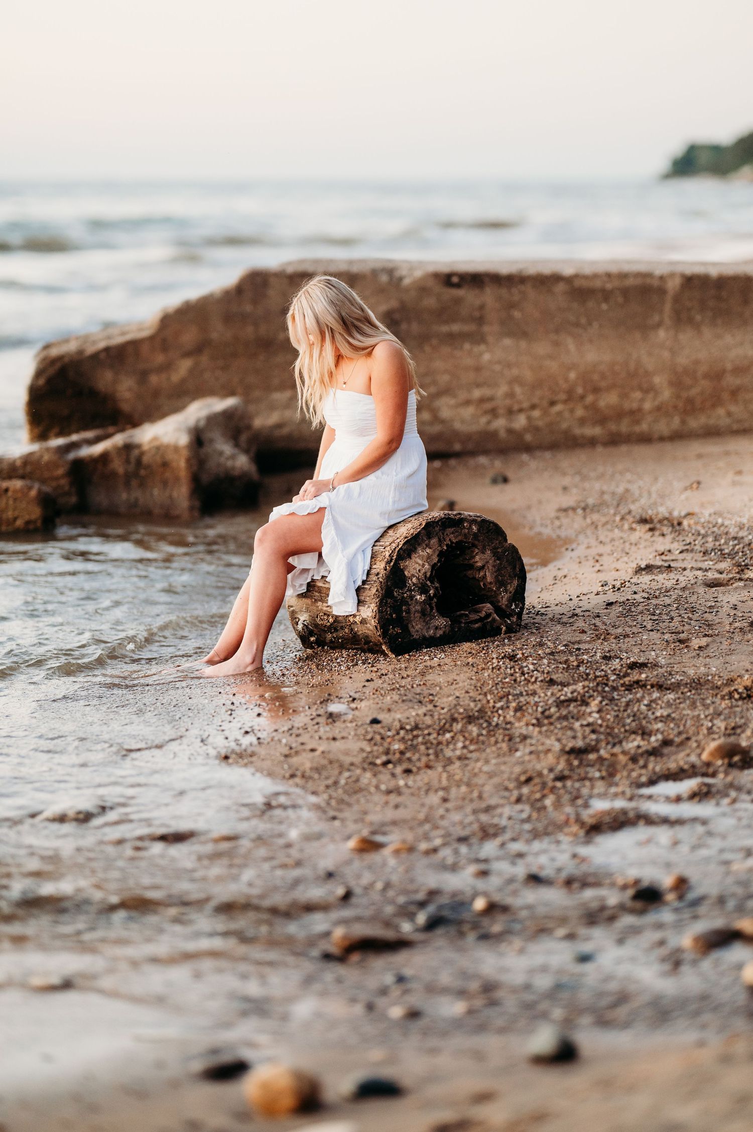 Dreamy coastal scene with person in white dress sitting on rocky beach at sunset near gentle ocean waves.