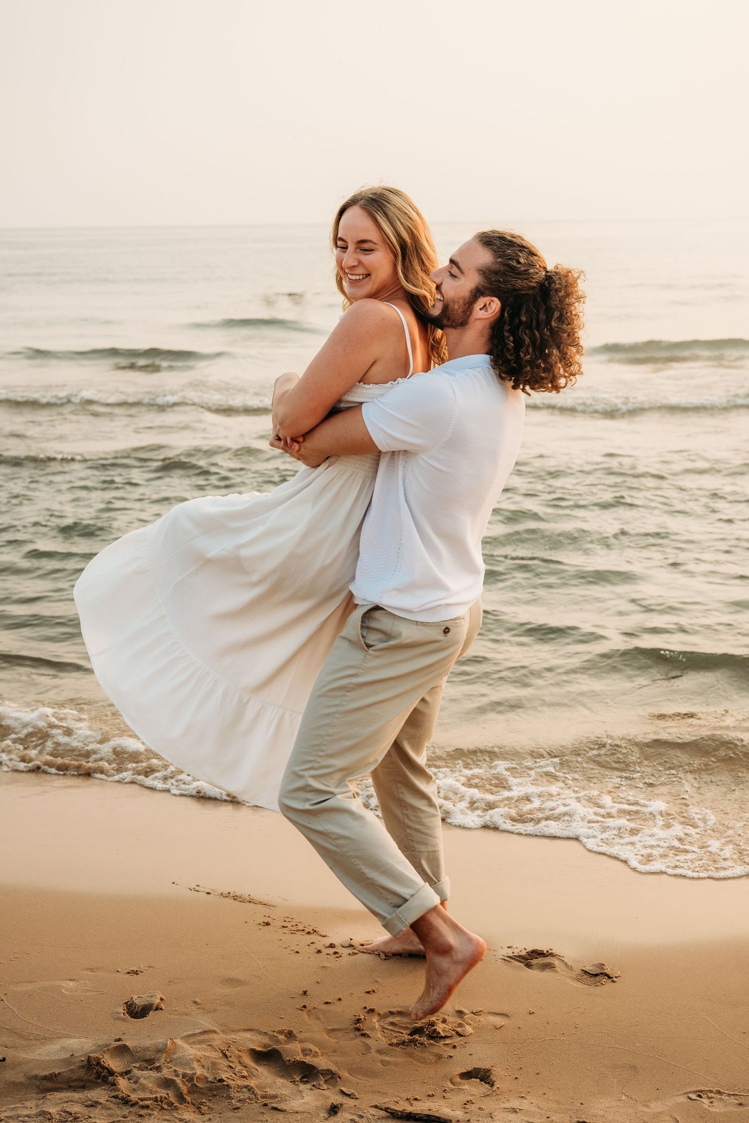 Playful couple in white outfits embracing and laughing together on a sandy beach at sunset.