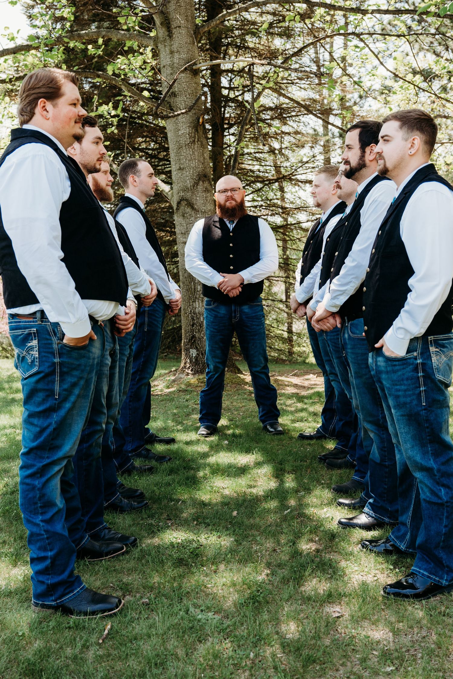 A groomsmen party in matching black vests and jeans stands in formation on grass beneath tall trees.