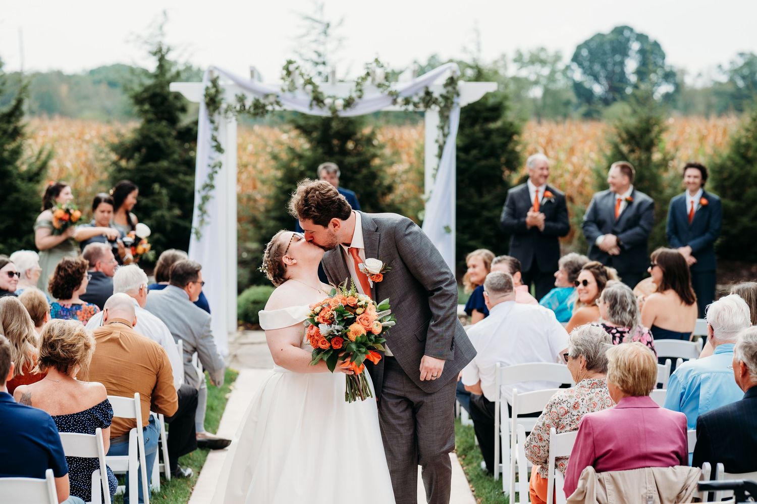 Outdoor wedding ceremony with guests seated in white chairs surrounded by fall foliage.