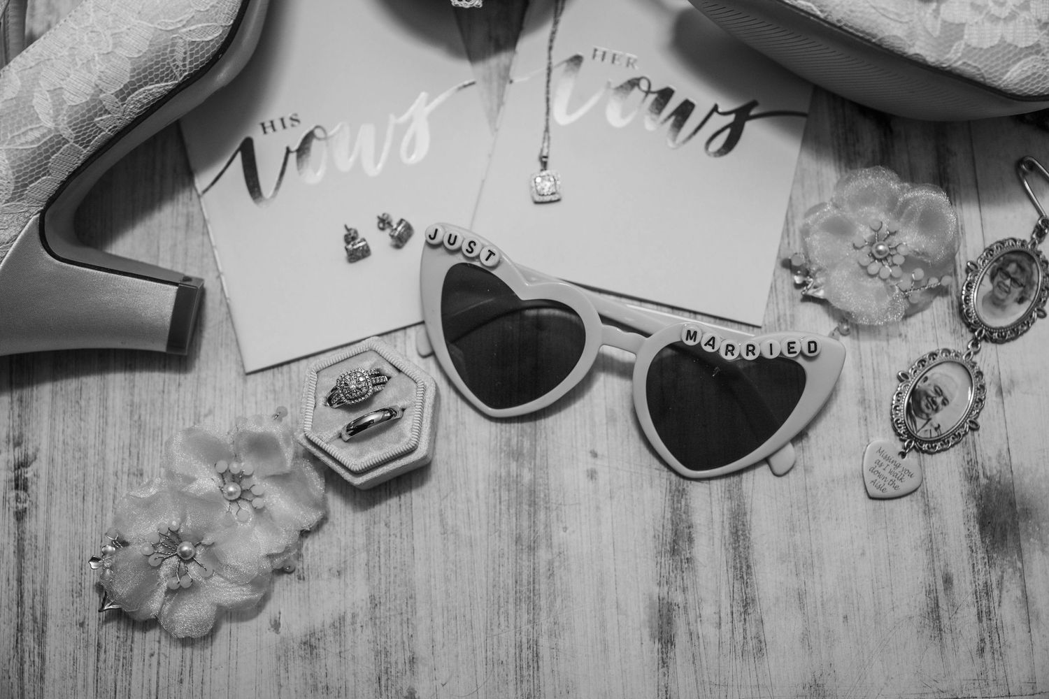 Black and white photo of heart-shaped sunglasses and wedding accessories on a wooden surface.