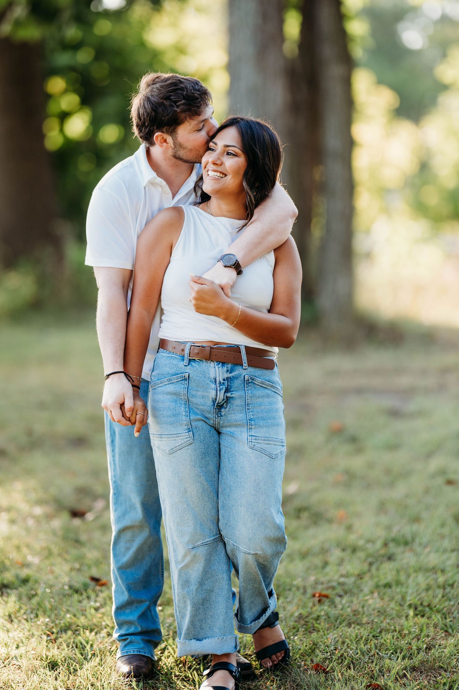 A couple wearing matching white tops and blue jeans embrace lovingly on grassy ground in soft natural lighting.