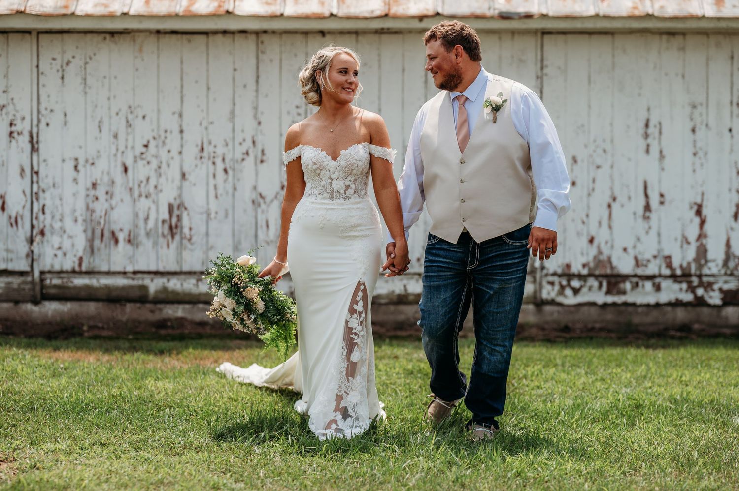 A couple sits together on grass against a rustic white barn wall during a romantic wedding photoshoot.