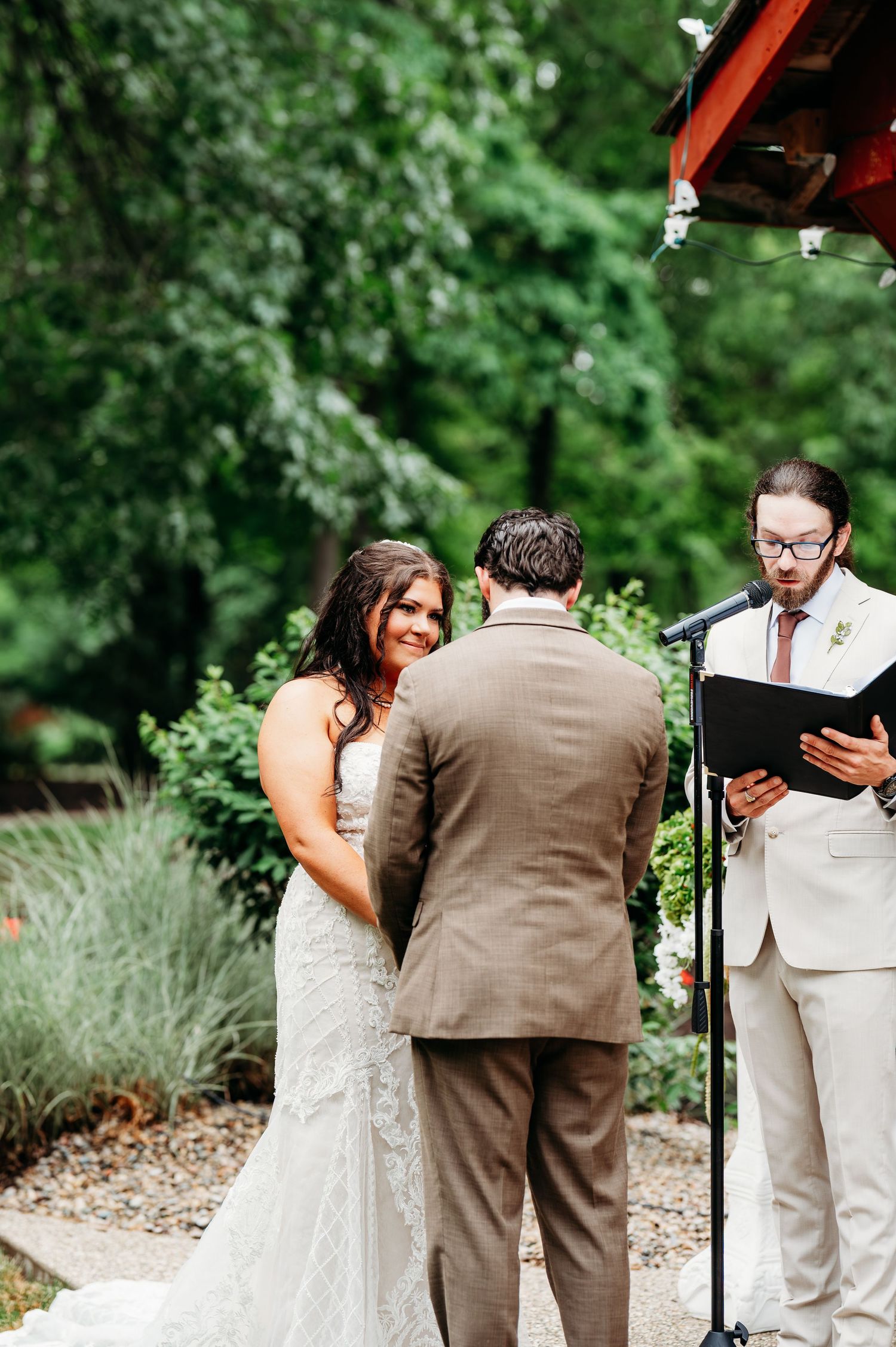 Bride smiling at groom during outdoor wedding ceremony while officiant reads from black book in lush garden.