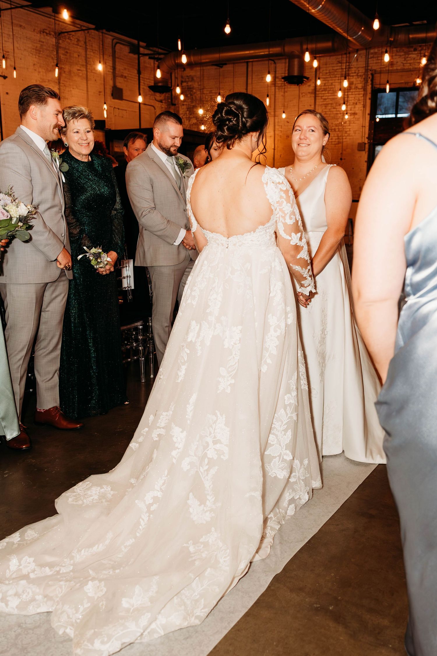 Wedding reception guests mingle in rustic barn venue with string lights while bride's lace train trails behind her.