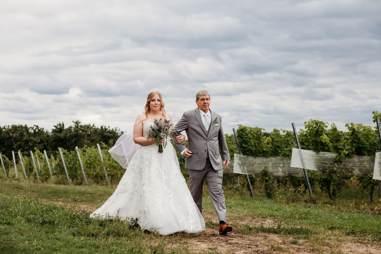 A couple walks arm in arm through a scenic vineyard on their wedding day under cloudy skies.