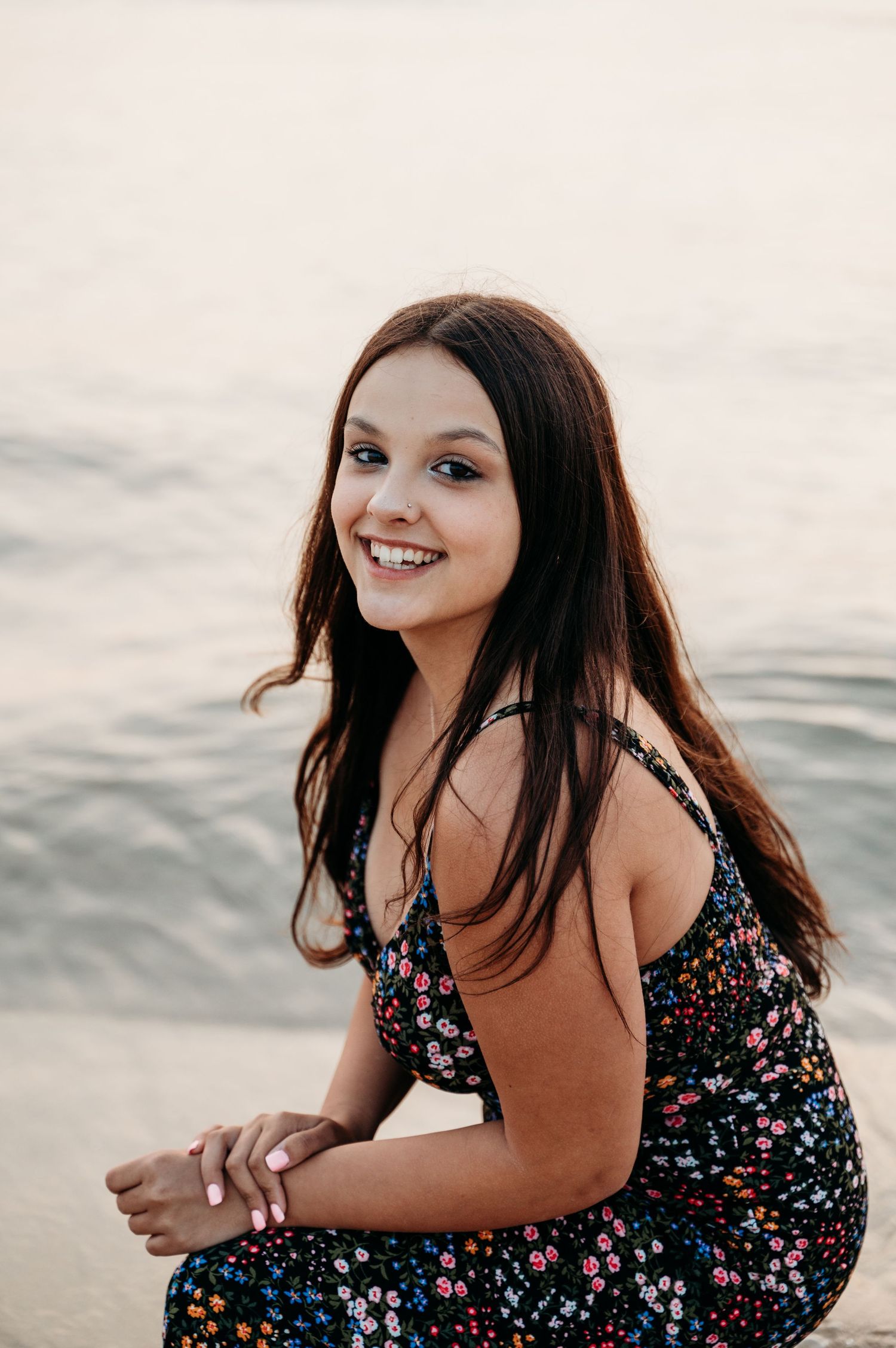 Young woman sitting by the water in a floral dress and wearing a bright smile on a calm evening.
